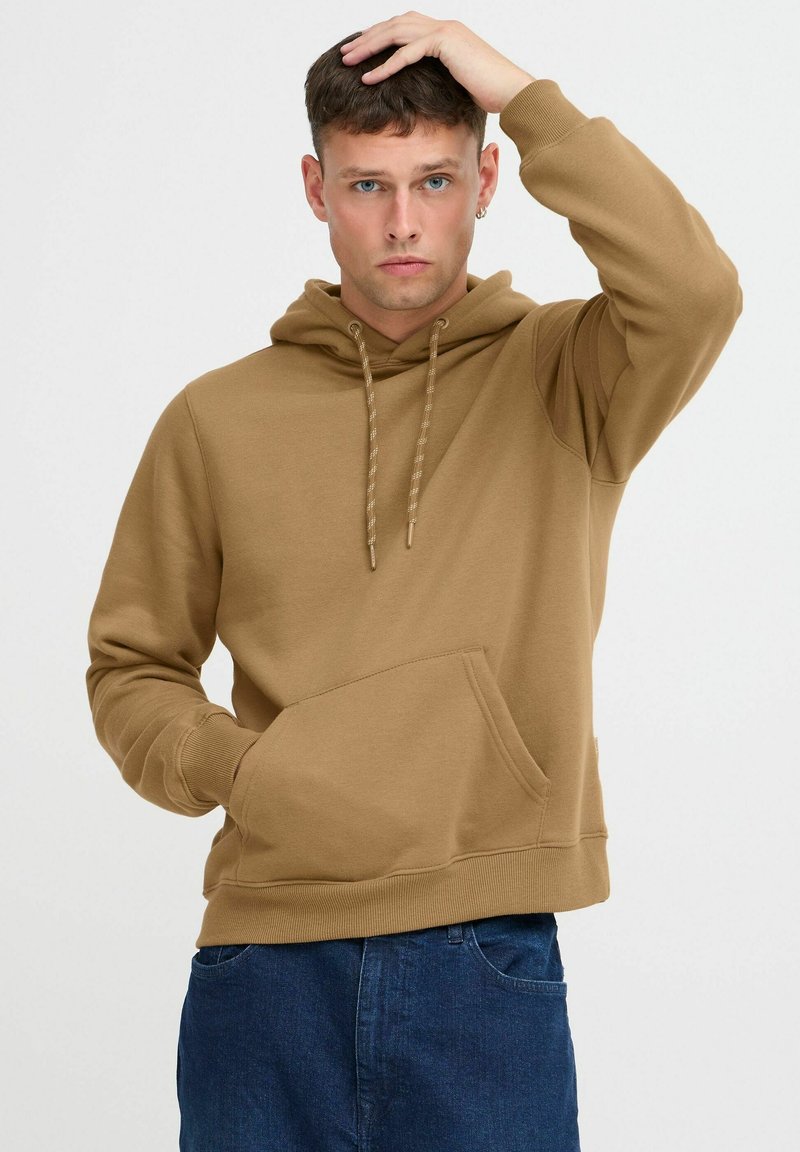 Brown hoodie made of cotton blend, featuring a front pocket, drawstring hood, and ribbed cuffs. Worn with dark blue jeans.