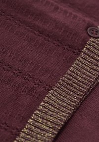 Burgundy stickad plagg med texturerat mönster och guldfärgad metalltråd som accent. Har en knappdetalj på vänster sida.