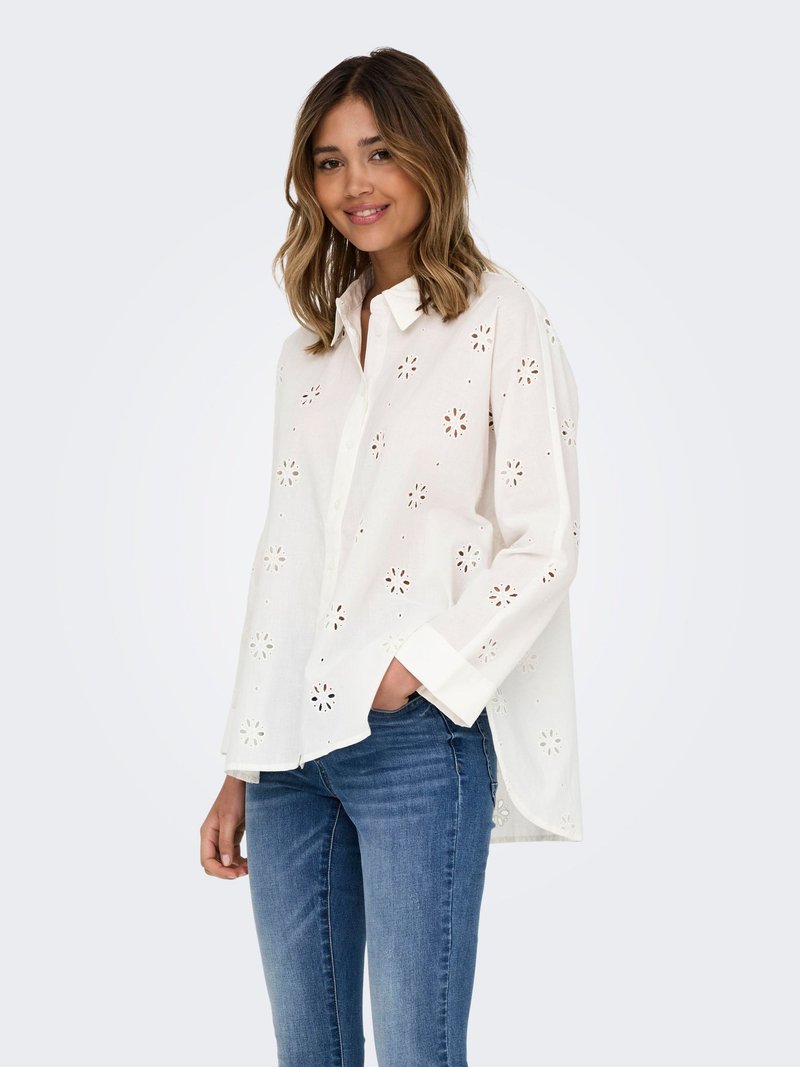 Tânără zâmbind, purtând o cămașă albă cu mânecă lungă și decupaje florale și blugi albaștri, cu mâna în buzunar, pe un fundal simplu.