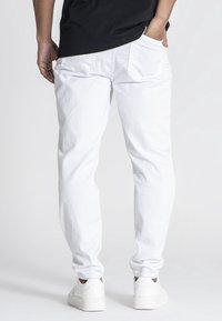 Pantalones vaqueros blancos con un corte cónico, que cuentan con bolsillos traseros y costuras sutiles. Combinados con una camisa negra y zapatillas blancas.