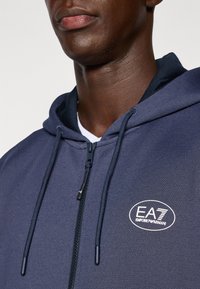 Sweatshirt com capuz e fecho éclair em azul-marinho, com um tecido texturizado, detalhes em cordão e um logotipo EA7 branco no peito.
