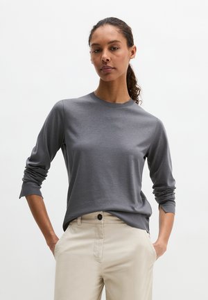 Kobieta z kręconymi włosami związanymi do tyłu, ubrana w szarą koszulę z długim rękawem i beżowe spodnie, stojąca z rękami w kieszeniach na białym tle.