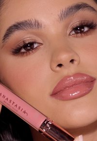Anastasia Beverly Hills flüssiger Lippenstift in einem sanften rosa Tube mit metallischen Akzenten, gehalten neben den vollen Lippen eines Models mit glänzendem Finish.