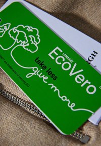 Grön EcoVero™-kort med vit text, trädgrafik och frasen "ta mindre, ge mer", delvis vilande på en strukturerad beige yta.