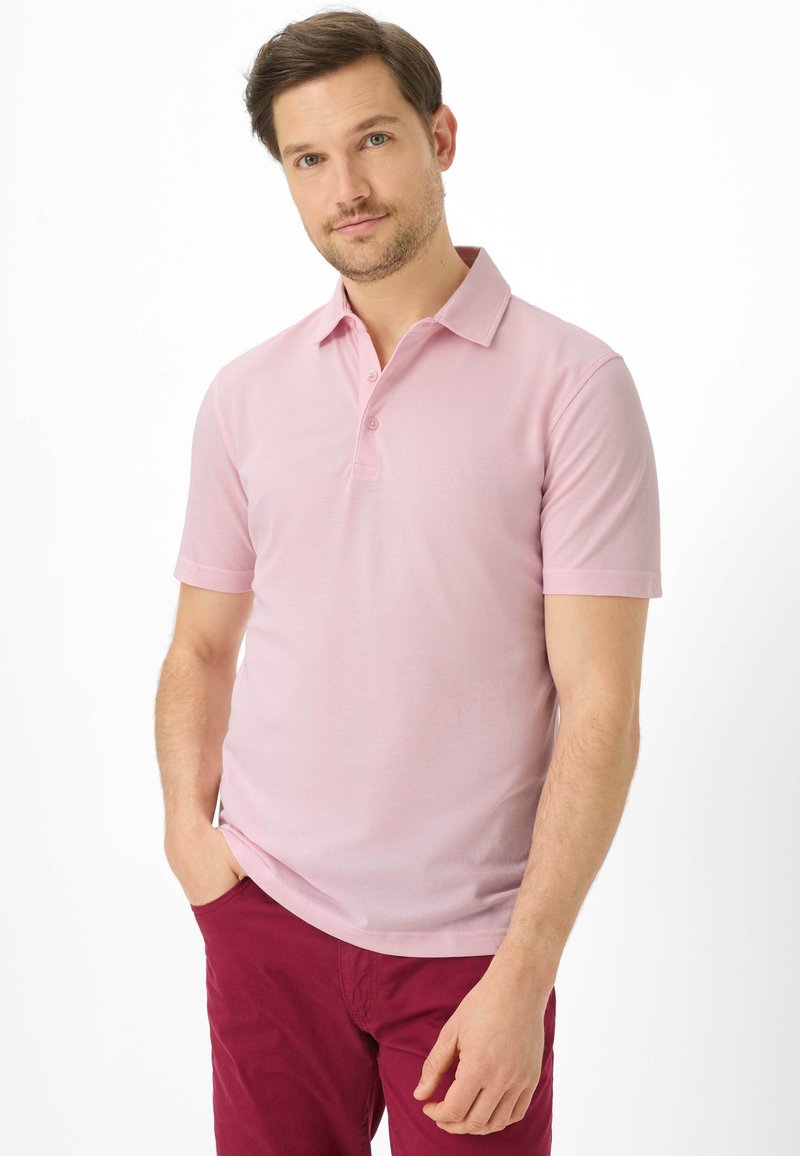 Homme aux cheveux bruns courts et légère barbe, portant un polo rose clair et un pantalon rouge foncé, debout avec une main dans la poche.