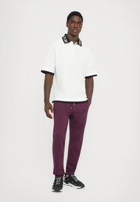 Camisa polo branca com colarinho preto, apresentando o texto "FUTURE", combinada com calças jogger roxas e ténis pretos. Design simples e casual.