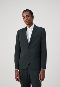 Dark green blazer with a notched lapel, single-button closure, and two front pockets, paired with a white collared shirt underneath.