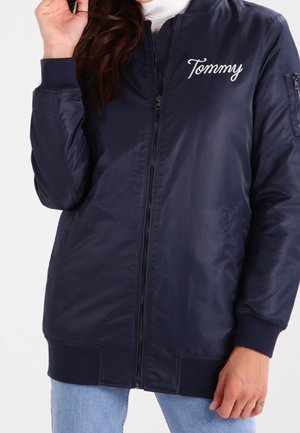 Blouson aviateur bleu marine avec fermeture éclair à l'avant et "Tommy" brodé sur la poitrine, porté sur un haut blanc et un jean bleu clair.