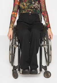 Un fauteuil roulant noir avec des roues argentées, placé devant une personne portant un haut à manches longues à fleurs et un pantalon noir.