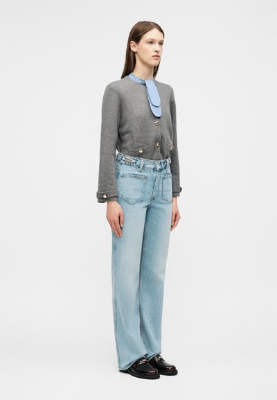 Γυναίκα που στέκεται, φοράει γκρι ζακέτα με κουμπιά και πλαϊνές τσέπες, ριγέ μπλε γραβάτα, ανοιχτό μπλε τζιν ψηλόμεσο και μαύρα μοκασίνια.