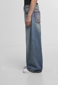 Osoba w luźnych, niebieskich dżinsach z czerwonymi szwami na kieszeniach i brzegach, w połączeniu z białymi trampkami, stojąca na jednolitym tle.