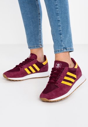 Osoba nosząca bordowo-żółte buty sportowe Adidas w połączeniu z podwiniętymi jasnoniebieskimi dżinsami, stojąca na białej powierzchni.