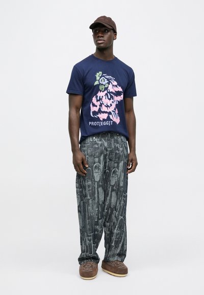 Μπλε ναυτικού τύπου μπλούζα με γραφικό σχέδιο που περιλαμβάνει λουλουδένια στοιχεία και κείμενο «PROTEGGIT». Φαρδιά γκρι παντελόνια με σχέδια, καφέ παντόφλες και καπέλο.