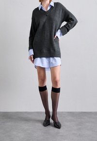 Maglione oversize grigio scuro con scollo a V, indossato sopra una camicia button-up azzurra chiara. Calze nere fino al ginocchio e ballerine nere appuntite completano il look.