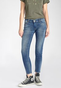 Jean skinny en denim bleu avec une finition décolorée, taille haute, et cinq poches, associés à des baskets montantes noires et un t-shirt vert olive.