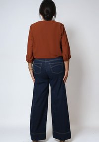 Chemise orange brûlée à manches 3/4, avec des accents de boutons, associée à un jean large bleu foncé avec des coutures renforcées et des poches arrière.