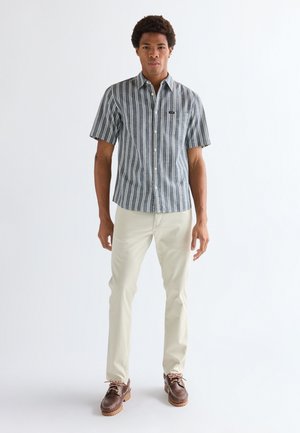 Jauns vīrietis stāv, valkājot zilu un baltu vertikālu svītru īsroku kreklu, gaišas bēšas bikses un brūnas auklām kurpes.