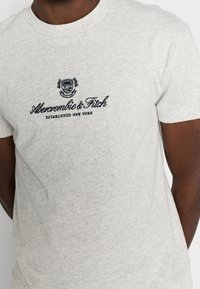 Camiseta de algodón gris con cuello redondo, que presenta un logo bordado en azul marino que dice "Abercrombie & Fitch Established New York" en el pecho.