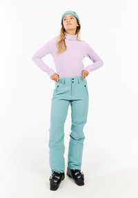 Un pull à col roulé côtelé violet clair associé à un pantalon de neige teal, présentant une taille haute, deux poches avant et des fermetures éclair noires, porté avec des bottes de ski noires.