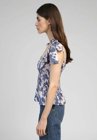 Haut à motif floral bleu et blanc, avec des liens aux épaules et un corsage smocké. Ourlet évasé. Porté avec un jean bleu clair.