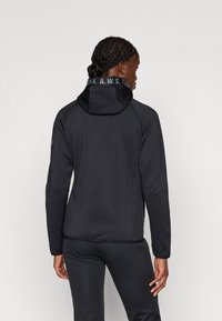 Mustat urheiluvaatteet, joissa on huppu, sileäpintaiset ja heijastavat logot kauluksessa; kapeasti istuva leikkaus, minimalistinen muotoilu.