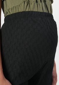 Pantalons noirs texturés avec une taille élastique, présentant un motif subtil et une coupe floue, associés à un haut vert olive.