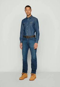 Mėlynas džinsinis marškinis su aštriu pečių iškarpu, dviem krūtinės kišenėmis, derinamas su vidutinio plovimo džinsais ir tan spalvos darbo batais. Modelis stovi neutralioje fone.