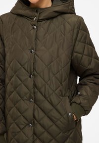 Veste matelassée olive avec capuche, dotée de boutons pression noirs, de poches latérales et de bords côtelés. Tissu texturé avec un motif en losanges.