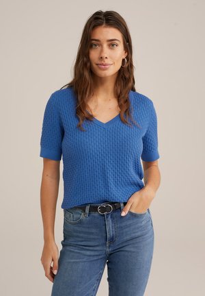 Blauer kurzärmeliger Strickpullover mit V-Ausschnitt, strukturiertem Stoff und gerippten Bündchen; kombiniert mit hoch geschnittenen blauen Jeans und einem schwarzen Gürtel.