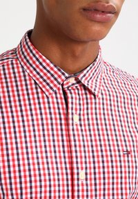 Camisa a cuadros rojos y blancos con cuello abotonado, que presenta un pequeño logo de acento azul marino en el pecho y botones de color claro.