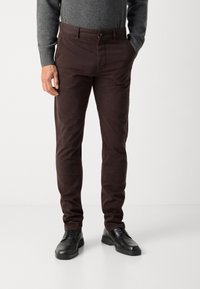 Brown cotton trousers with a slim fit, featuring a button closure and front pockets. Worn with black lace-up shoes and a gray sweater.