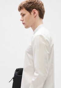 Λευκό πουκάμισο με κουμπιά και όρθιο γιακά, με λεία υφή. Μαύρη τσάντα με φερμουάρ που κρατιέται στο πλάι. Μινιμαλιστικός σχεδιασμός, ουδέτερο φόντο.