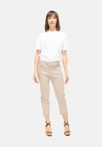 Donna in piedi di fronte che indossa una camicia bianca a maniche corte, pantaloni beige con bottoni e risvolti, e sandali neri con cinturini.
