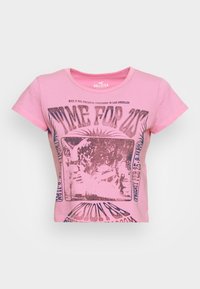 Camiseta de algodón rosa con cuello redondo y mangas cortas, que presenta una impresión gráfica en morado oscuro, mostrando texto e imágenes en el centro.