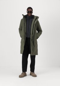 Olivgrüne Parka mit Kapuze über einer schwarzen Zip-Jacke, kombiniert mit schwarzen Hosen und hellbraunen Schnürboots, die eine elegante, strukturierte Oberfläche aufweisen.