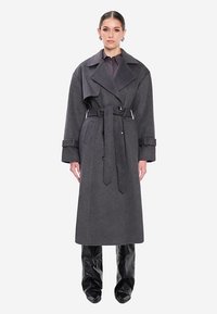 Grauer Wollmantel mit Gürtel in der Taille, breiten Revers und Seitentaschen. Kombiniert mit glänzenden schwarzen Hosen. Zeitloses, maßgeschneidertes Design.