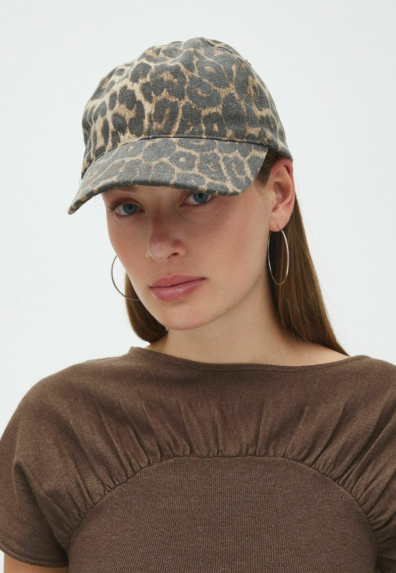 Femme aux longs cheveux bruns portant un haut froncé marron, de grandes boucles d'oreilles et une casquette de baseball imprimé léopard.