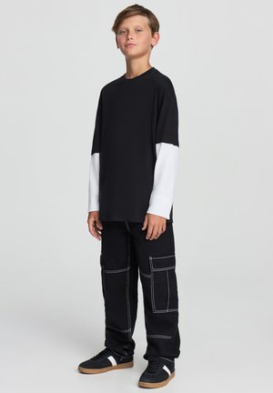 T-shirt nera con maniche lunghe bianche, abbinata a pantaloni cargo neri con cuciture bianche e molteplici tasche. Sneakers nere con dettagli bianchi.