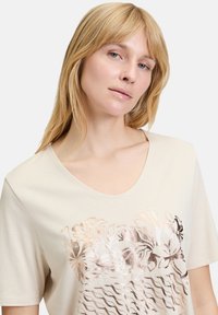 Creme kortærmet t-shirt med v-hals, med metalisk blomsterprint og bølgete linjemønstre på forsiden. Blødt, glat bomuldsstof.