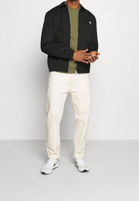 Homme portant une veste noire zippée, une chemise vert olive, un pantalon crème et des baskets blanches, debout les mains jointes devant lui.