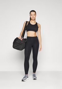 Soutien-gorge de sport noir avec logo, leggings noirs taille haute, baskets grises et un sac de sport noir avec des accents texturés.