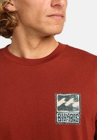 Hnědorudé tričko vyrobené z bavlny, se záplatou s designem vlny a textem "BILLABONG" černou barvou, obrysem s bílým okrajem.