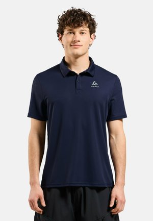 Junger Mann mit lockigem Haar, der ein marineblaues Poloshirt mit kurzen Ärmeln und einem kleinen Logo auf der linken Brust trägt.