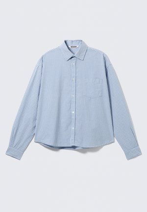 Svetlo modra srajca z dolgimi rokavi, gumbi spredaj, z majhnim karo vzorcem in žepom na prsih, položena na belo površino.