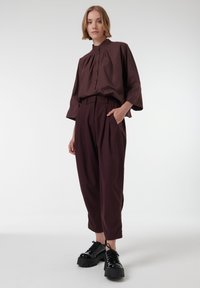 Blusa color marrón con un cuello alto y cierre de botones en la parte delantera, combinada con pantalones holgados marrones y zapatos de plataforma negros. Textura suave, ajuste relajado.