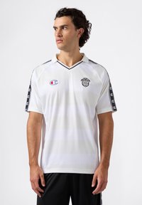 Jonge man met krullend donker haar, gekleed in een wit sportshirt met zwarte afwerking en logo's, kijkt naar links tegen een effen achtergrond.