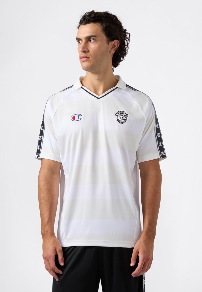 Jonge man met krullend donker haar, gekleed in een wit sportshirt met zwarte afwerking en logo's, kijkt naar links tegen een effen achtergrond.