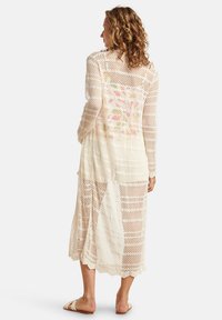 Langer, leichter Überwurf in Creme, mit einem Häkelmuster und offenen Geweben sowie gewellten Kanten. Texturiert und luftig.