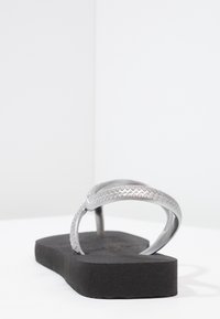 Hopeasaluiset hopeanväriset flip-flopit, joissa on teksturoitu hihna ja musta tasapohja. Hihnassa on sahalaitakuviota, joka on suunniteltu mukavuutta ja rentoa käyttöä varten.