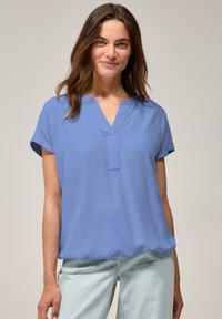 Blouse à manches courtes, bleu clair, avec un col en V, offrant une coupe décontractée et une texture douce ; se marie avec un pantalon en denim clair.